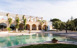 Uma área de piscina com pergolado, arcos de pedra e um edifício branco.