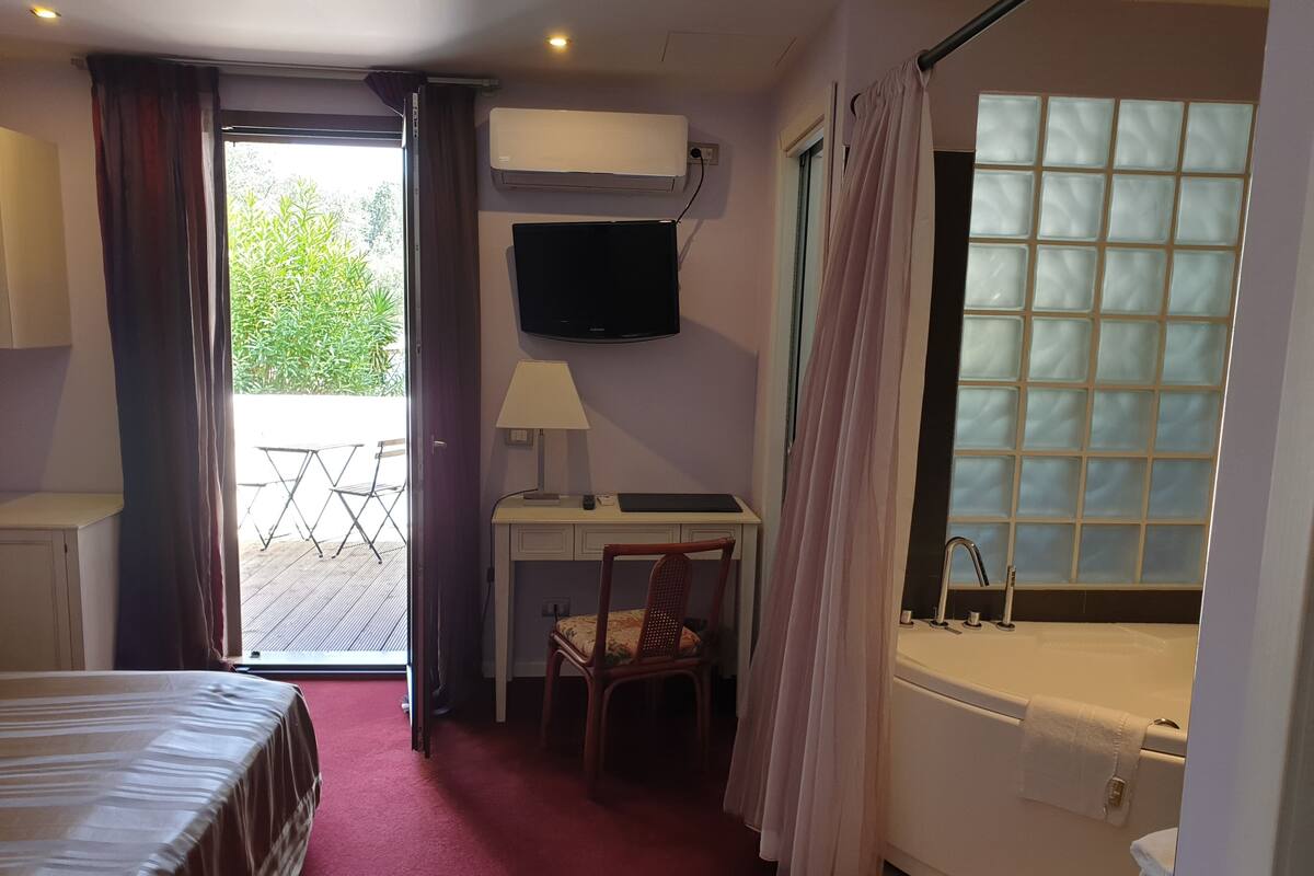 A hotel room with a bed, desk, chair, TV, and a bathroom with a bathtub and window.