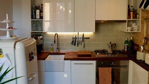 Una cocina compacta con muebles blancos, horno empotrado y pileta. Hay dos lámparas colgantes, un refrigerador y varios objetos de cocina sobre las encimeras.