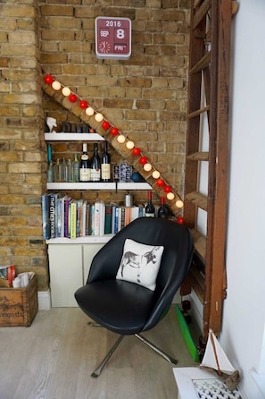 A cozy corner with a black chair, a bookshelf with books and wine bottles, a brick wall, and a date sign.