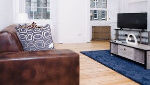 A modern living room with a brown leather sofa, patterned cushions, a blue rug, a wooden floor, and a television on a wooden stand. - Elforma - Barony Suite (Edinburgh)