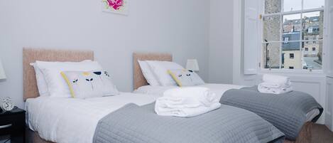 A double bed with a headboard, a bedside table with a lamp, and a window with a view of buildings.