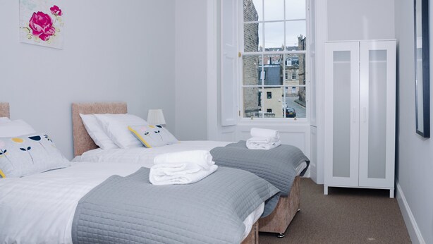 A hotel room with two beds, a window with a city view, a wardrobe, and a floral artwork on the wall. - Elforma - Barony Suite (Edinburgh)