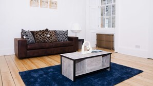 A living room with a brown sofa, patterned cushions, a wooden coffee table, a blue rug, and a window with a view of buildings. - Elforma - Barony Suite (Edinburgh)