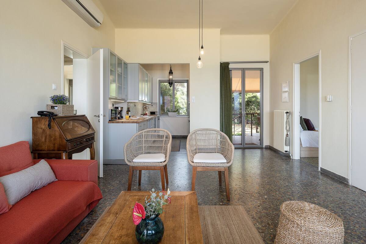 A spacious living area with a red sofa, wicker chairs, a coffee table with a vase, and a kitchen in the background.