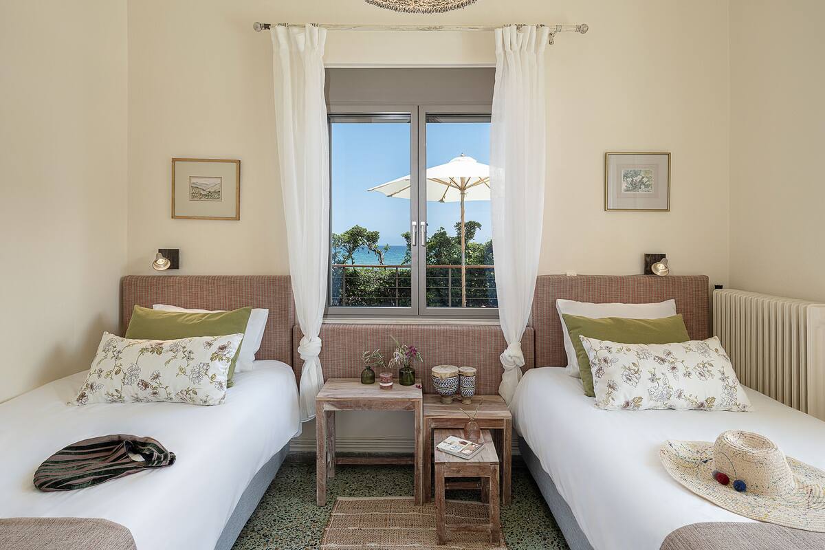 A bedroom with two beds, a small table, and a view of the sea through a window.