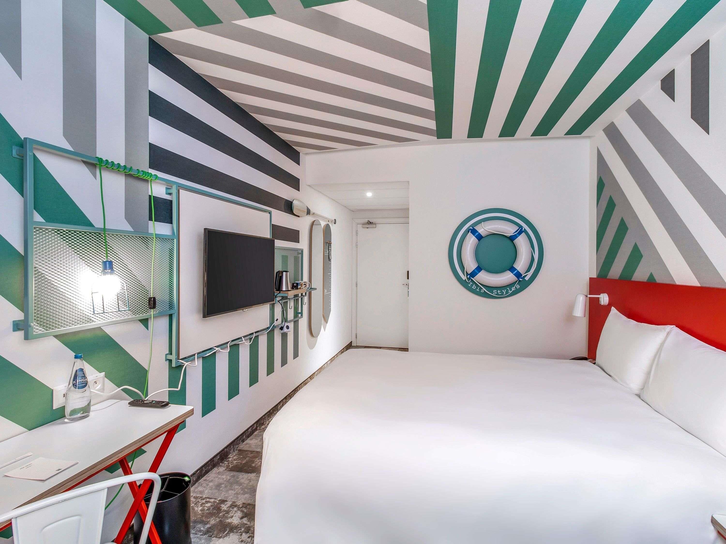A hotel room with a bed, a desk with a chair, a TV, a mirror, and a circular wall decoration.