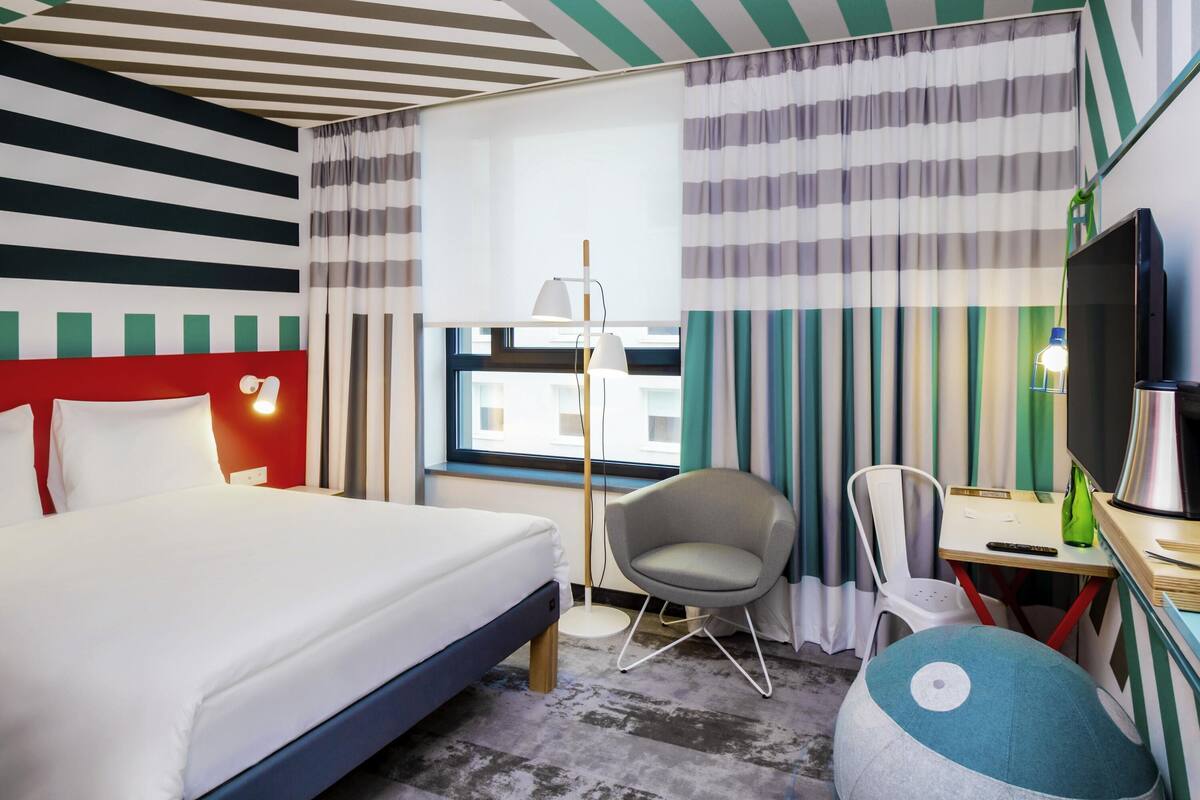 A hotel room with a bed, a desk, a chair, a TV, and a window with curtains.