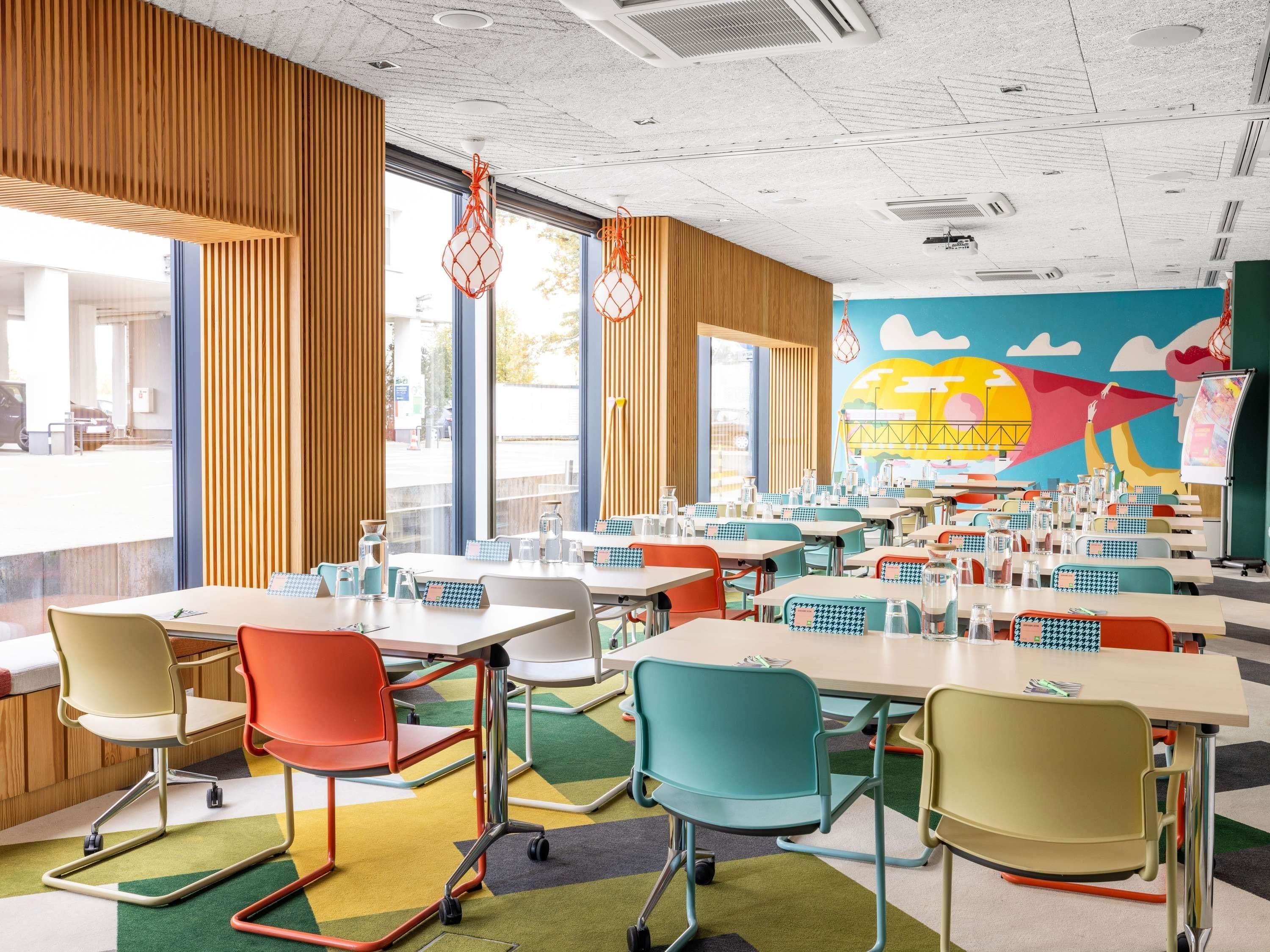 Eine hell erleuchtete Cafeteria mit bunten Stühlen und Tischen, großen Fenstern und einem Wandgemälde.