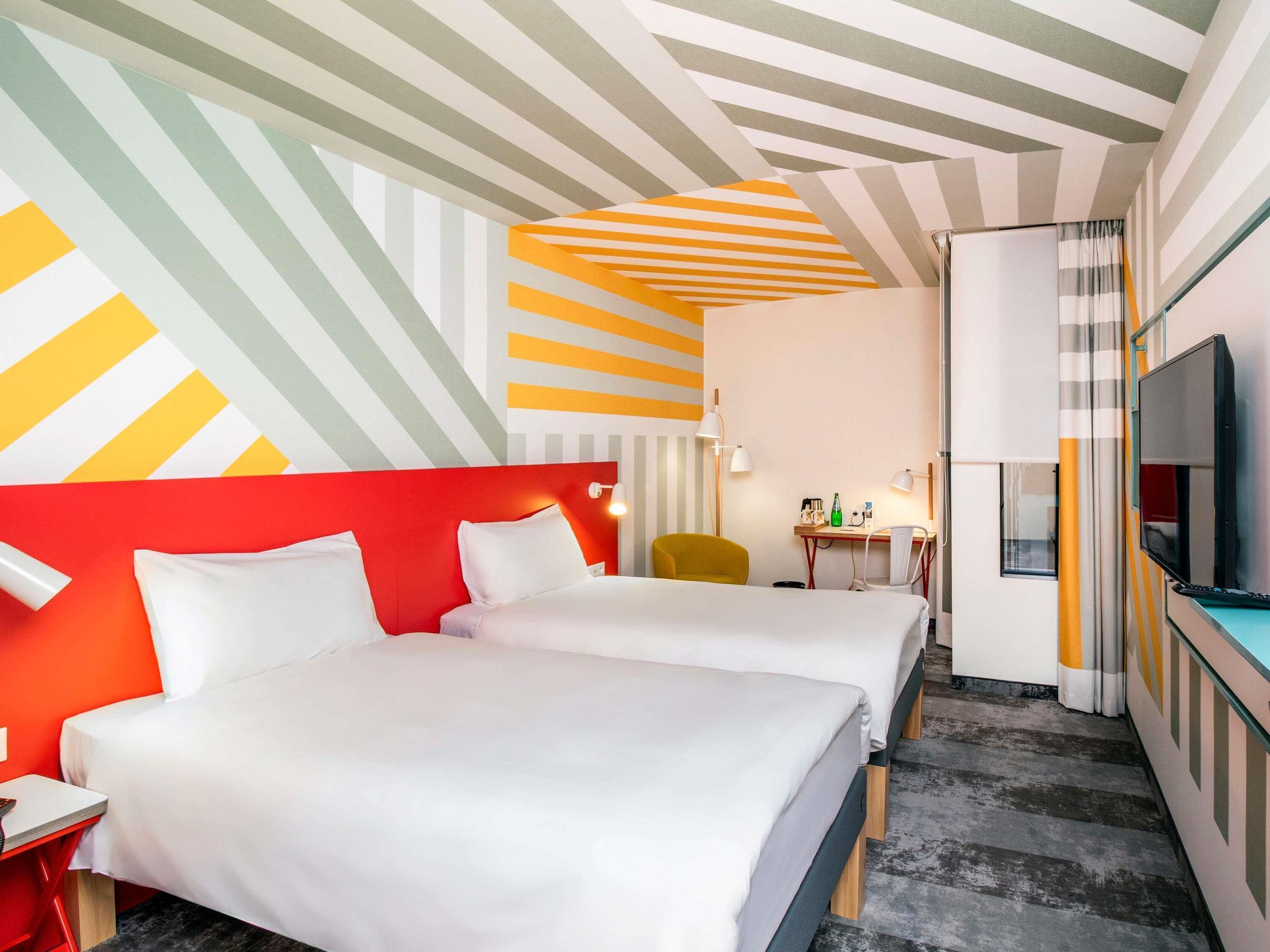 A hotel room with two beds, a desk, a chair, a TV, and a striped ceiling.