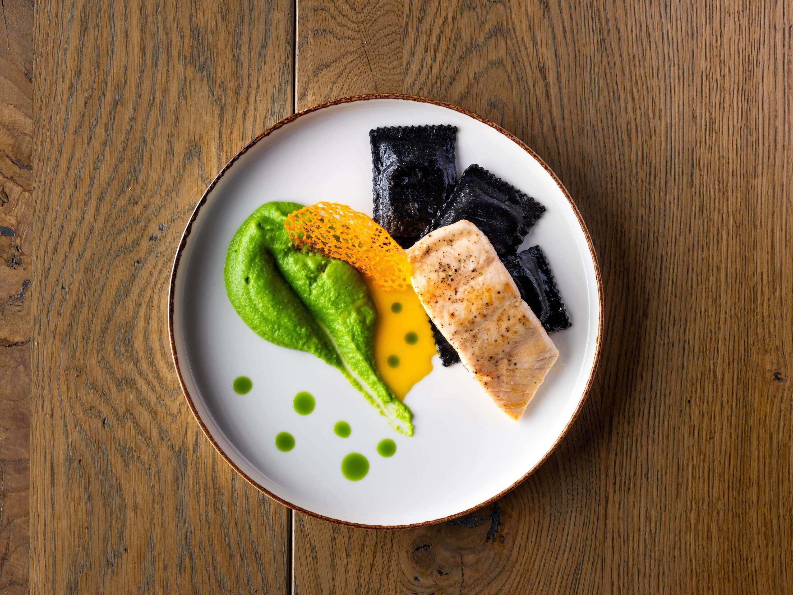 A plate with a piece of fish, green sauce, and black and yellow items on a wooden table.