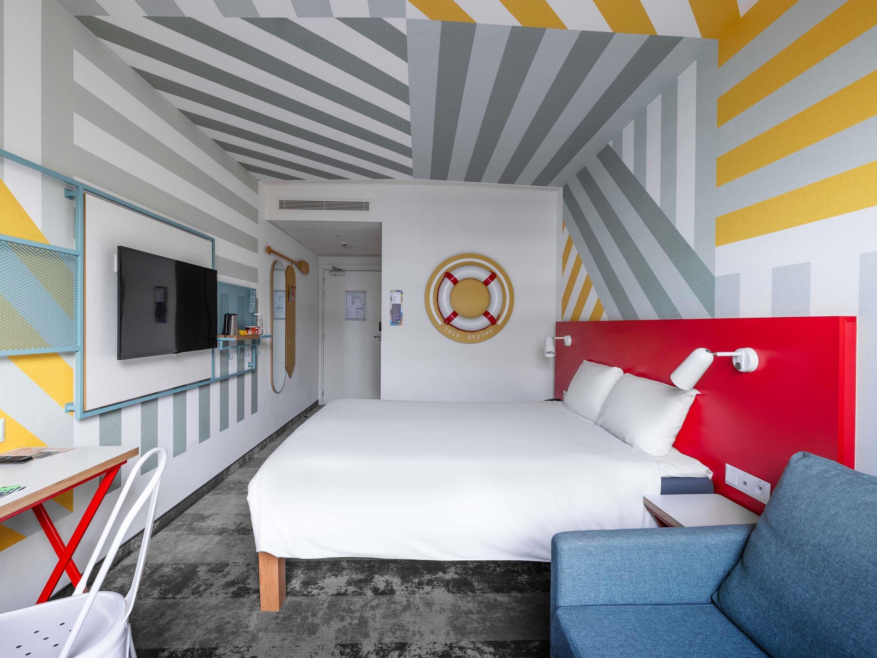 A modern hotel room with a bed, a desk with a computer, a TV, and a red accent wall.
