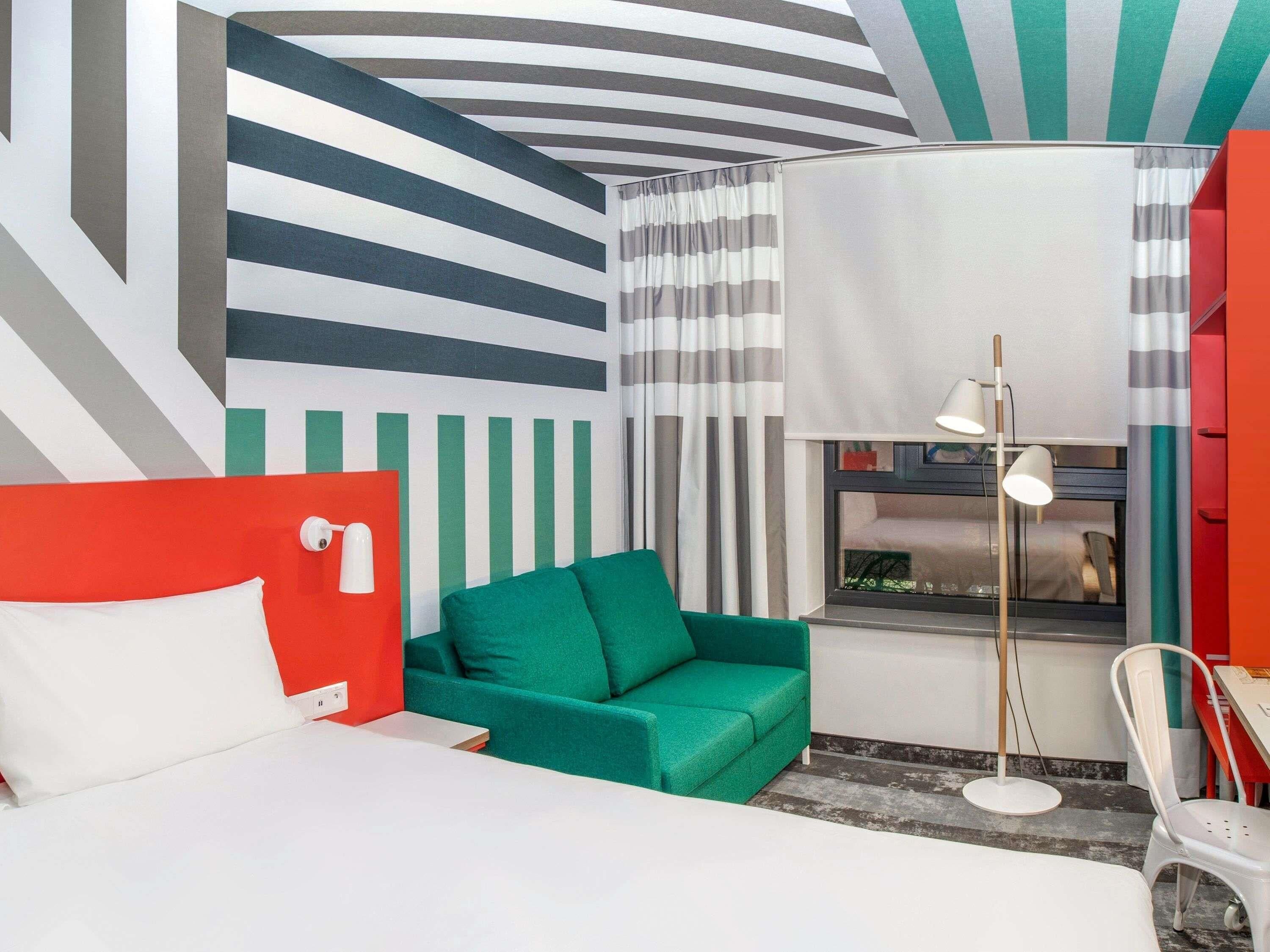A hotel room with a bed, a green sofa, a red accent wall, a window with curtains, and a floor lamp.