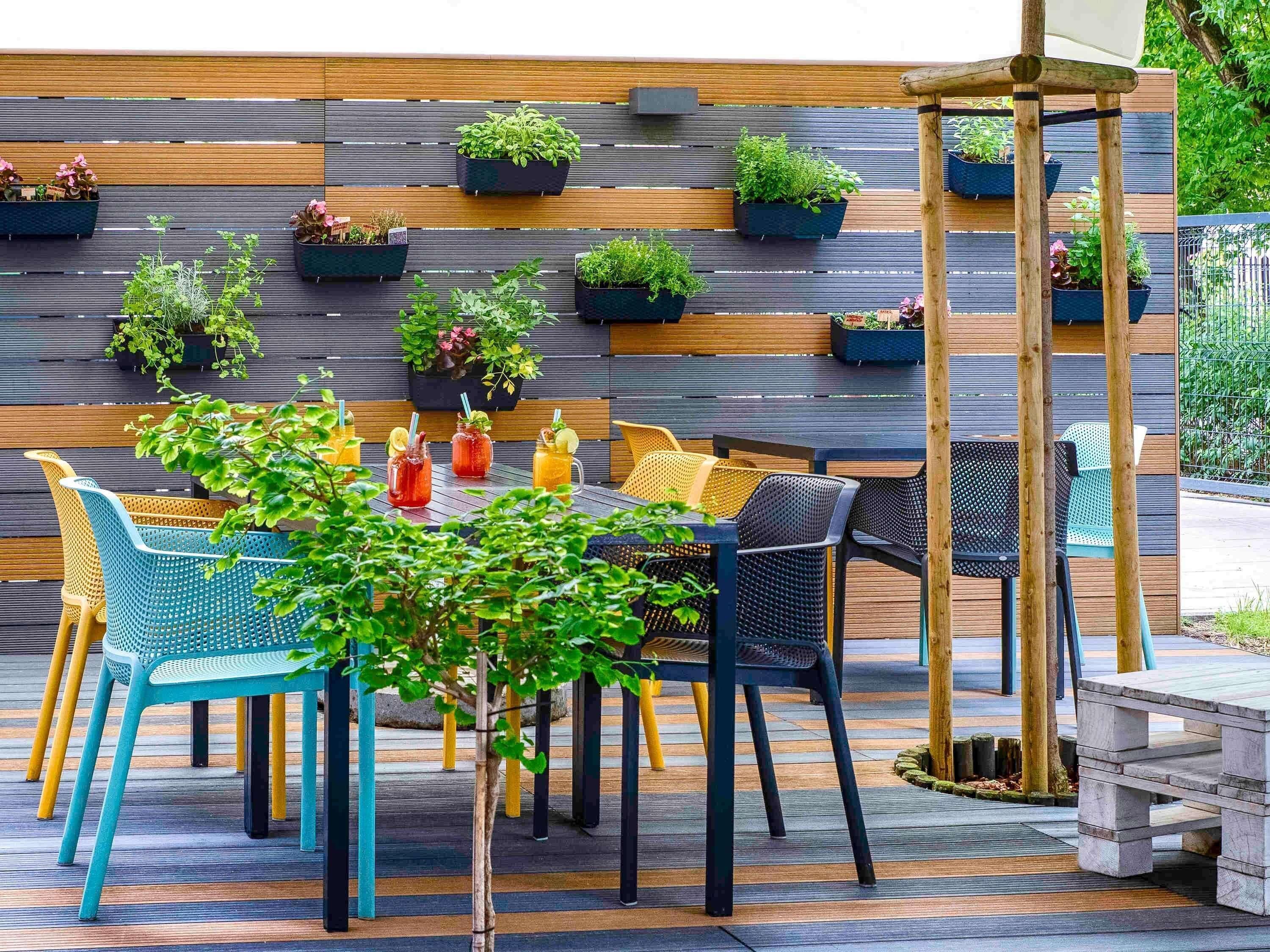 An outdoor seating area with a wooden wall and hanging planters, featuring a variety of plants and a table with chairs.