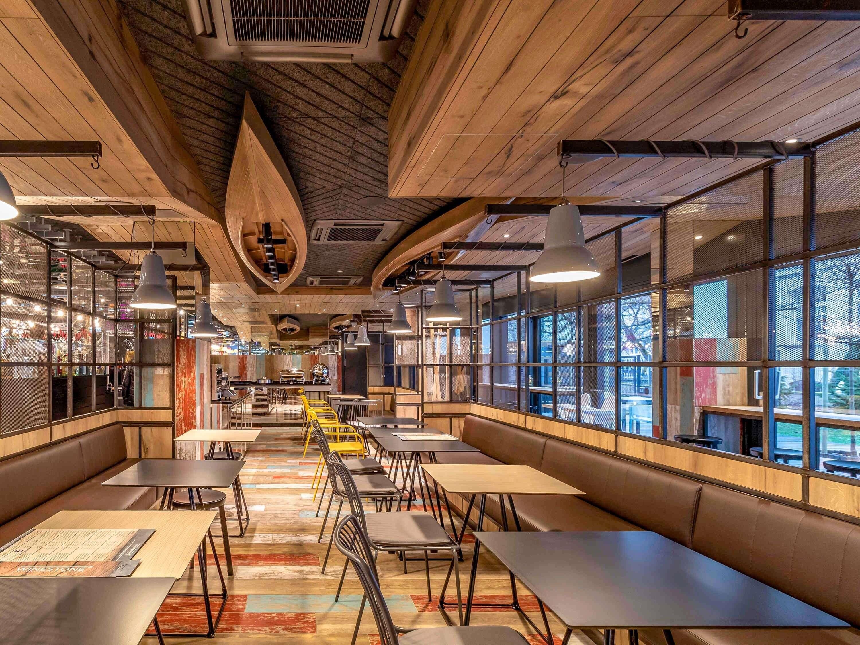 A restaurant interior with high ceilings, exposed beams, and a mix of natural and artificial lighting. Tables and chairs are arranged for dining.