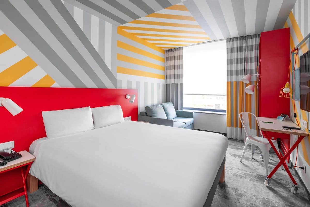 A hotel room with a large bed, a desk with a chair, a small sofa, a window with curtains, and a geometric-patterned ceiling.