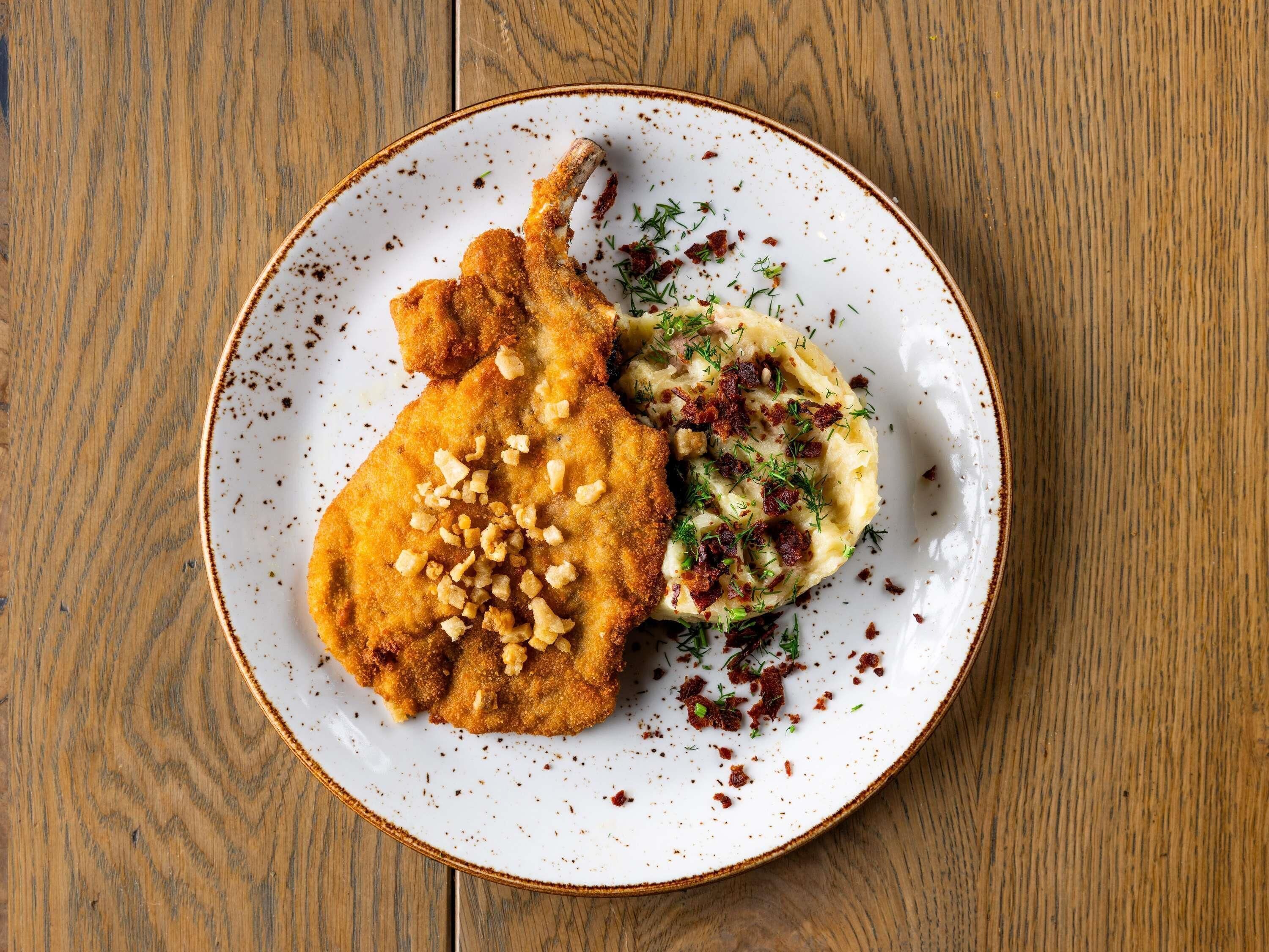 Un plato con un preparado empanizado y frito, posiblemente una milanesa de pollo, y puré de papas decorado con hierbas y trozos de tocino.