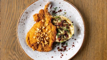 Un plato con un preparado empanizado y frito, posiblemente una milanesa de pollo, y puré de papas decorado con hierbas y trozos de tocino.