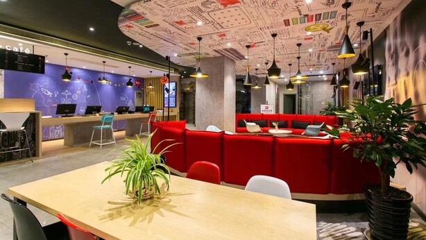 Een moderne hotellobby met een receptiebalie, een rode zithoek en een grote plant.