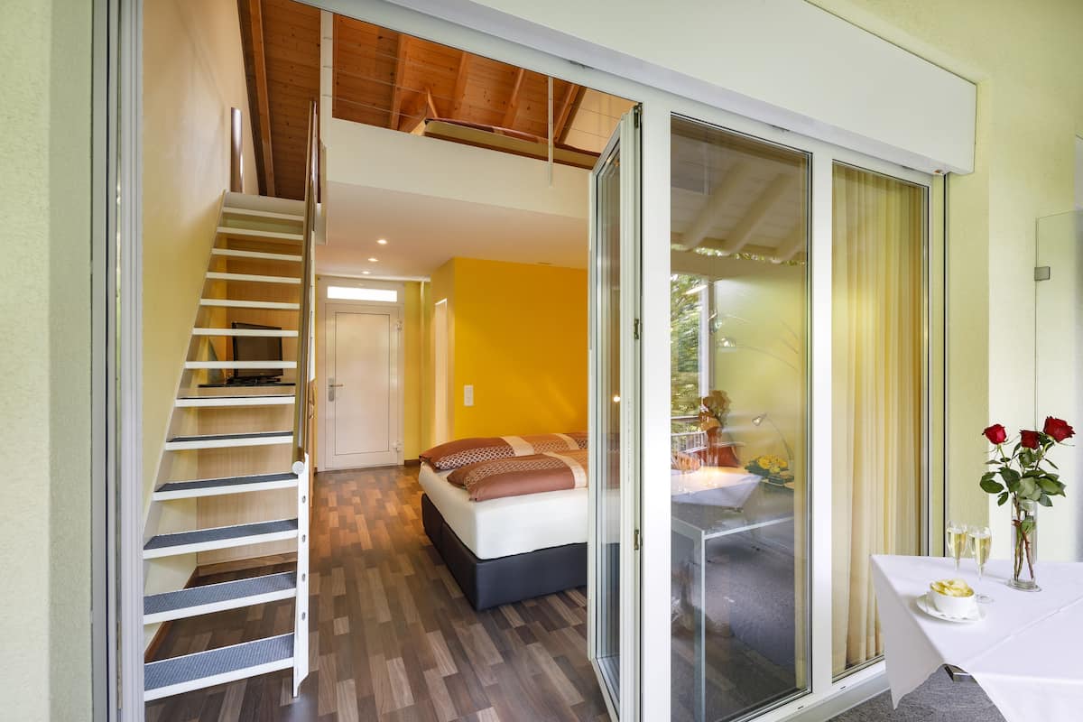 Une chambre d’hôtel moderne avec un escalier en bois, un lit doté d’une tête de lit sombre et un coin repas avec une table dressée pour deux personnes.