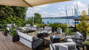 Un área de estar al aire libre con mobiliario moderno, plantas en macetas y vistas a barcos en un lago.