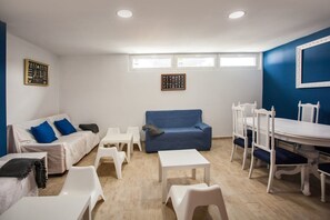 A room with a blue sofa, white chairs, a white table, and a dining table with chairs. There are two windows with blinds, and a framed picture on the wall.