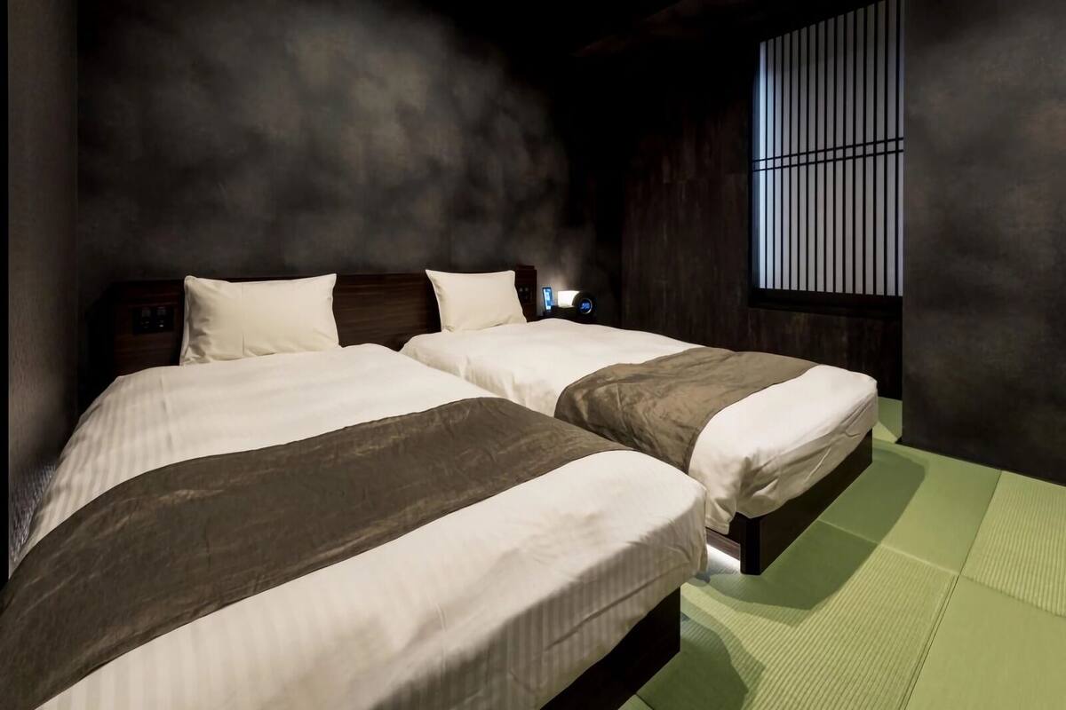 Una habitación con dos camas, una ventana con persianas verticales y un despertador fijado a la pared.