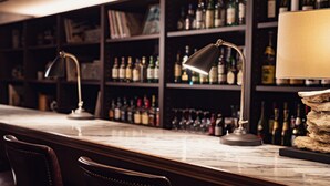 Zona de bar con encimeras de mármol, dos lámparas de barra regulables, sillas de cuero y estanterías repletas de botellas de licores.