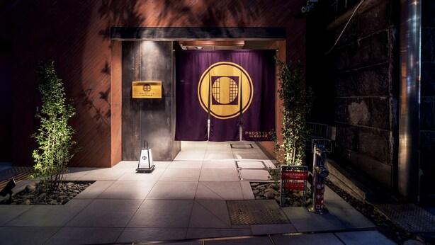 Entrada de un edificio con fachada de madera, un cartel púrpura con un emblema dorado y un pequeño árbol al frente.