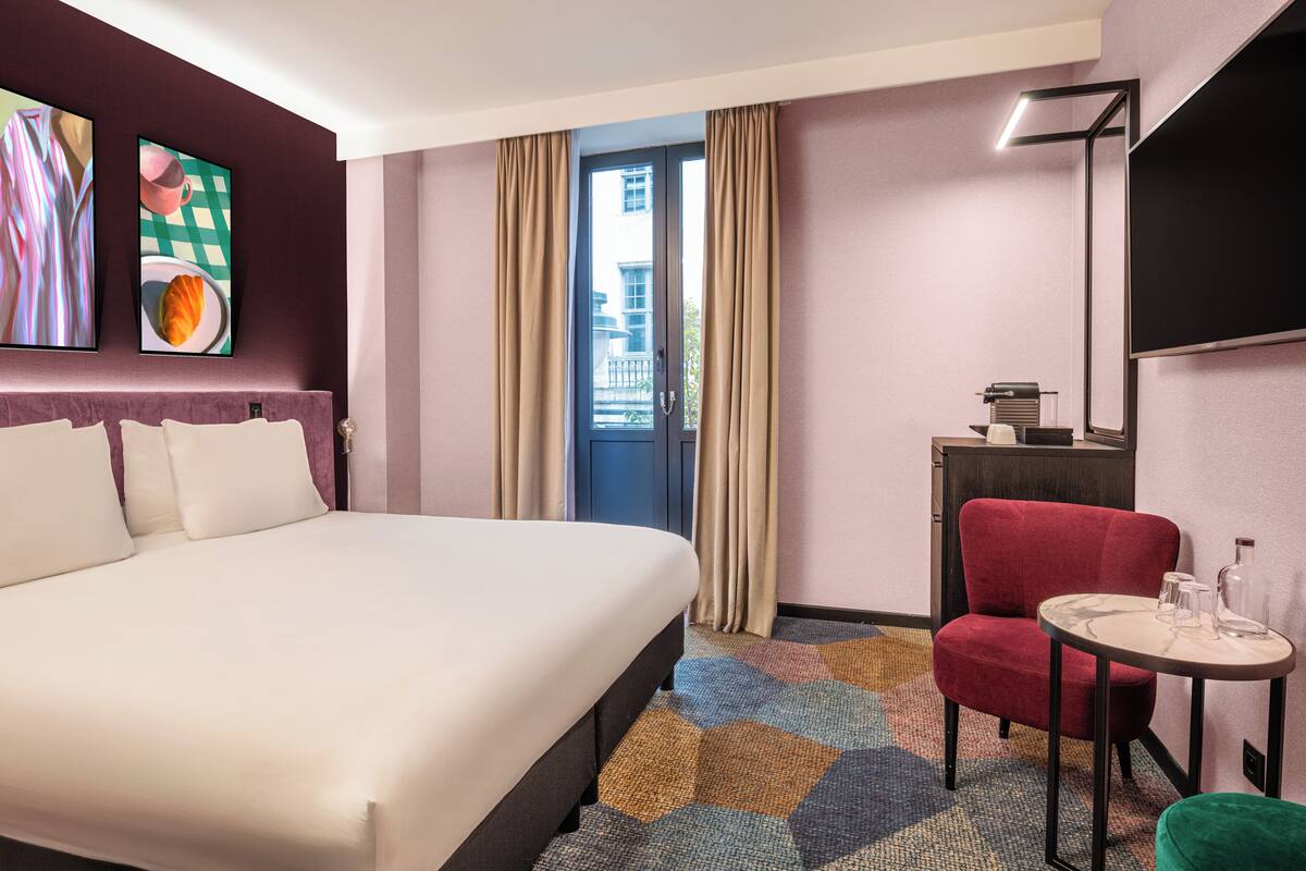 Habitación de hotel con cama, una silla roja, una mesita con vasos y un televisor en la pared.