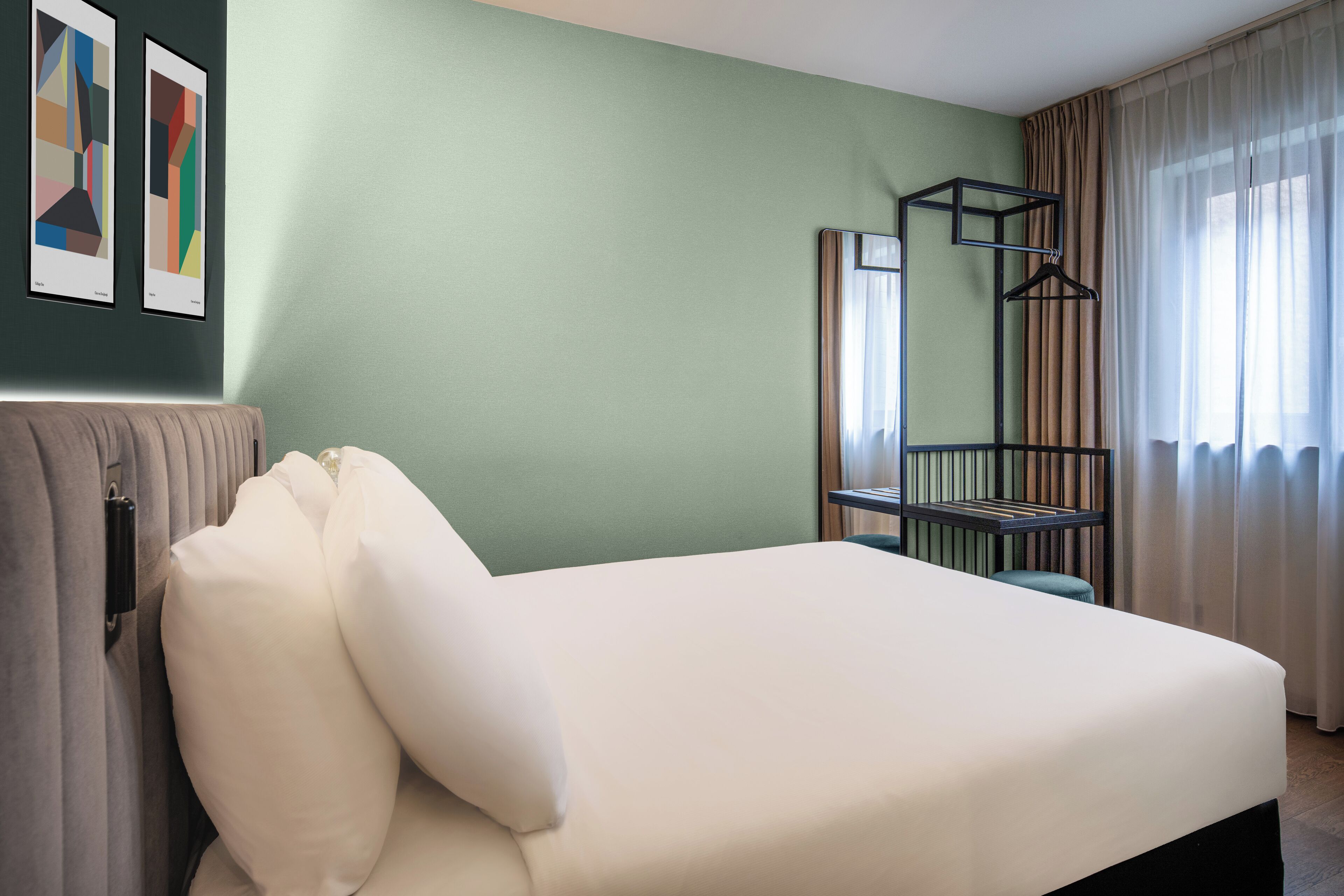 Habitación de hotel con cama, radiador, perchero y ventana con cortinas.