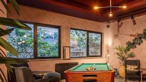 Una mesa de billar en una habitación con dos ventanas, un sofá, una silla y una planta.