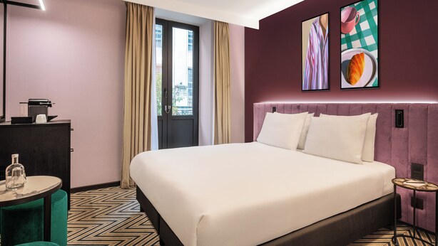 Una habitación de hotel con una cama grande, un escritorio con lámpara, una mesita redonda y dos cuadros enmarcados en la pared.