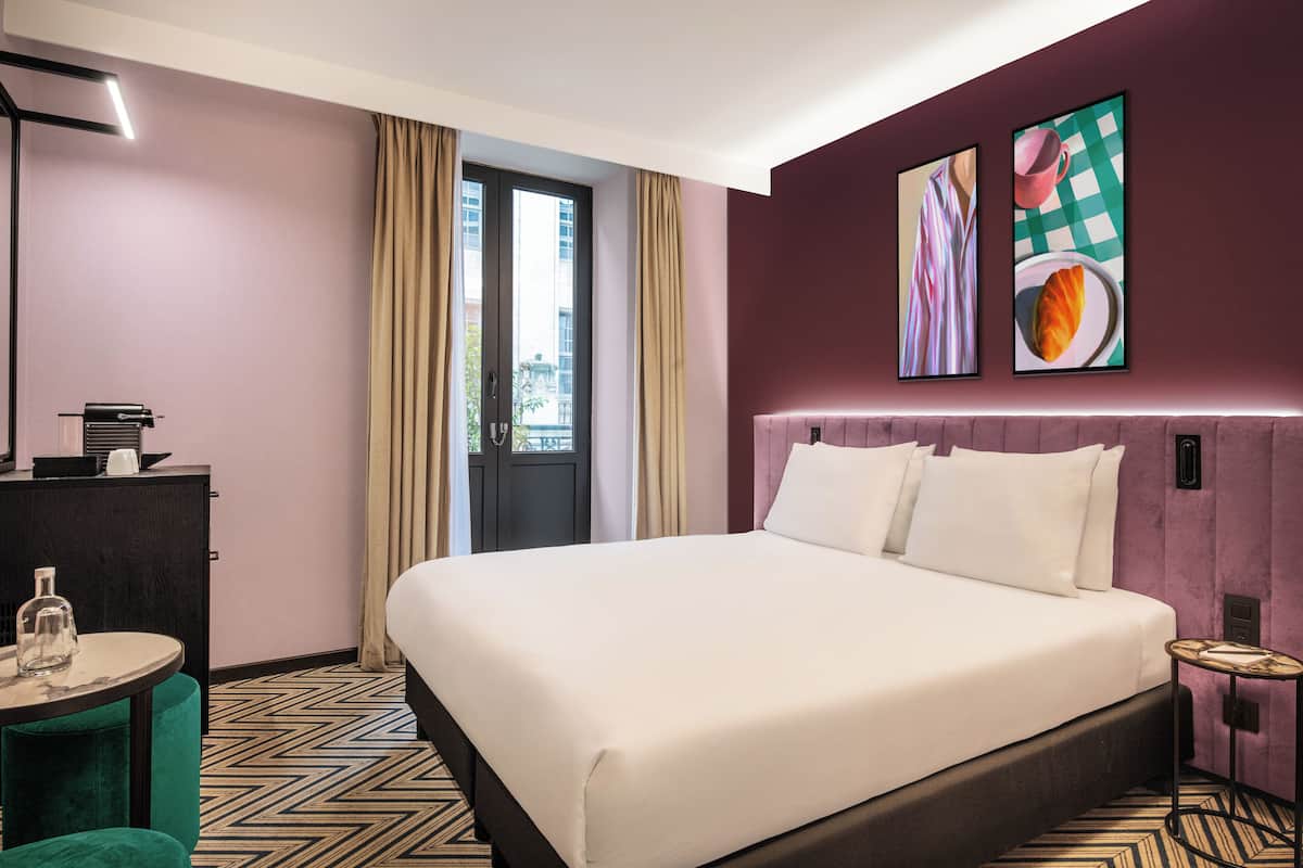 Una habitación de hotel con una cama grande, un escritorio con lámpara, una mesita redonda y dos cuadros enmarcados en la pared.