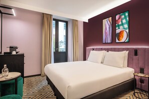 Une chambre d’hôtel avec un grand lit, un bureau avec une lampe, une petite table ronde et deux œuvres d’art encadrées accrochées au mur.