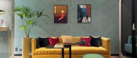 Een moderne woonkamer met een gele bank, groene poefjes, een laptop en twee ingelijste kunstwerken aan de muur.
