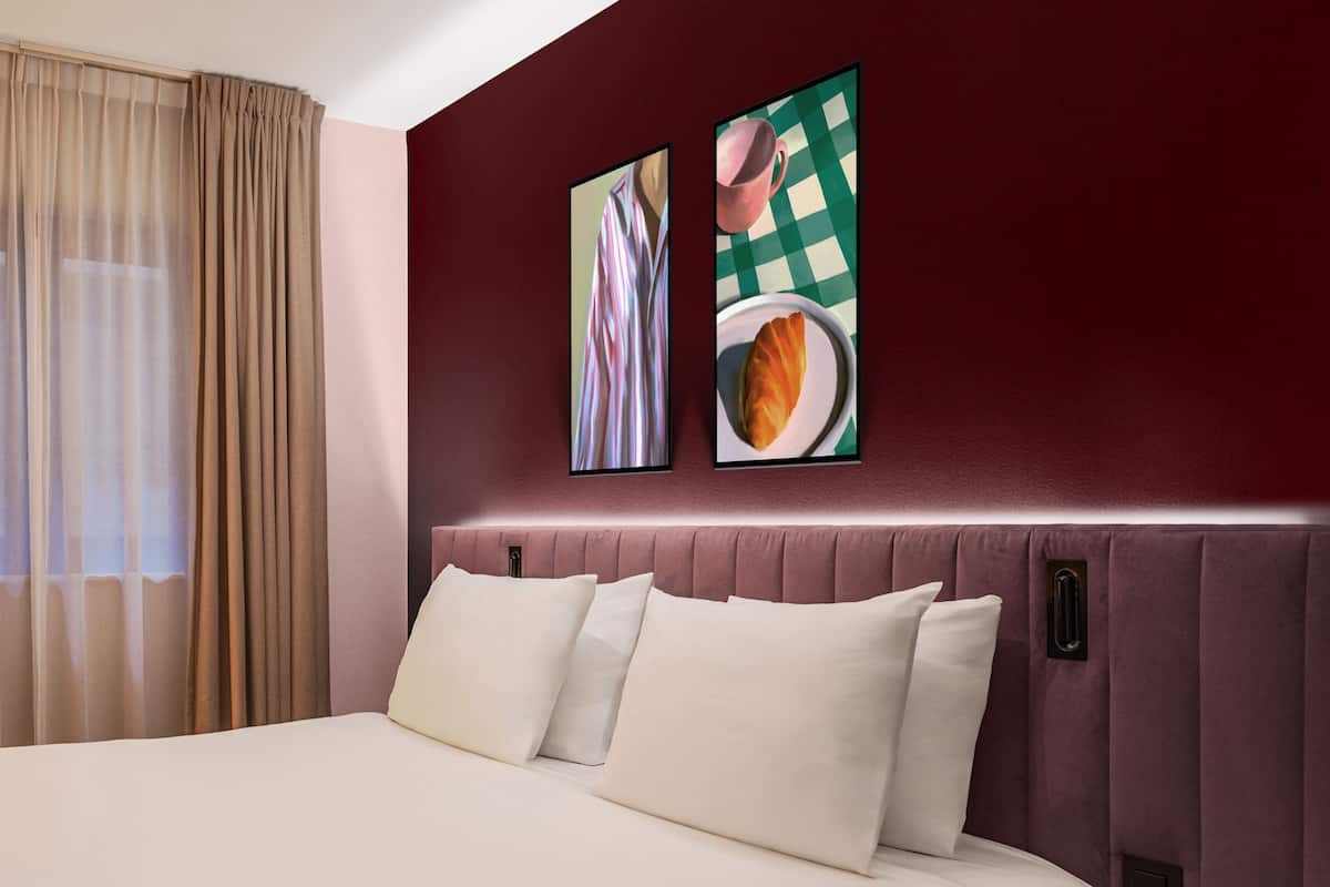Una habitación de hotel con una cama grande, ropa de cama color beige, un cabecero con un panel rosa claro y dos obras de arte enmarcadas en la pared.