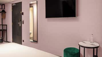 A hotel room with a bed, a television mounted on the wall, a mirror, a small table with a bottle and glass, and a green ottoman.