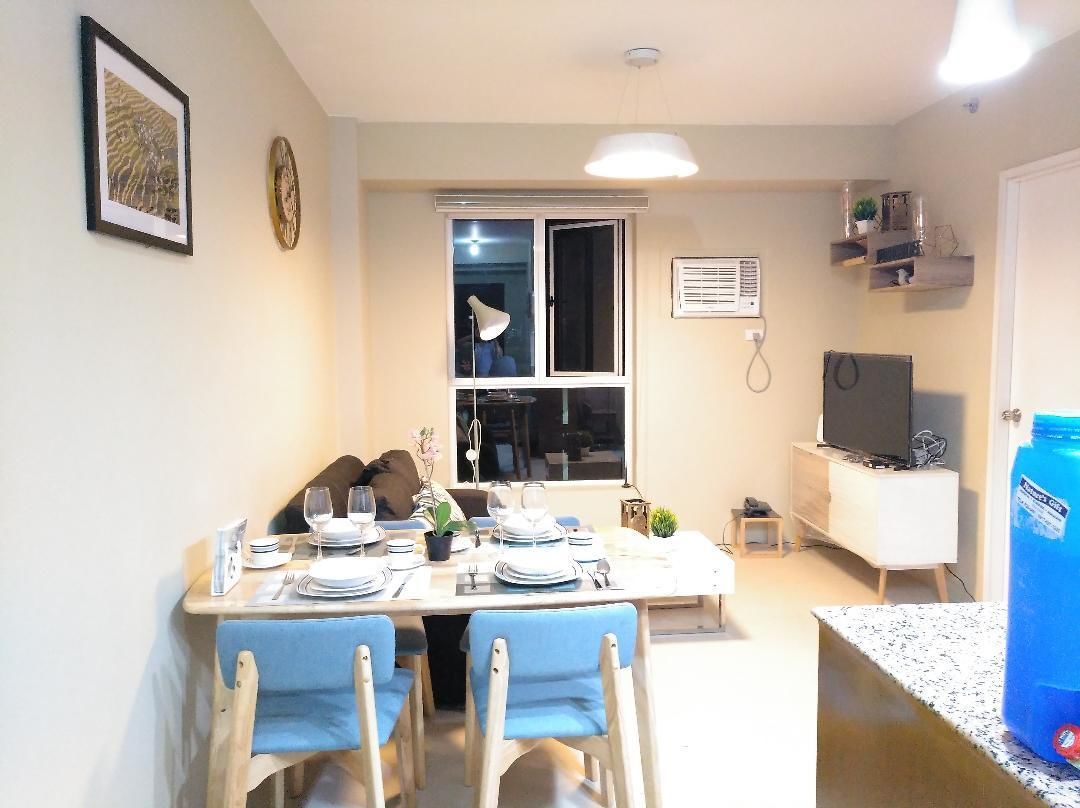 A dining area with a table set for four, a window with blinds, a television on a stand, and a water dispenser.