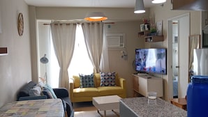 Una sala de estar con un sofá amarillo, una mesa de centro, un televisor y una cocina con un dispensador de agua azul.