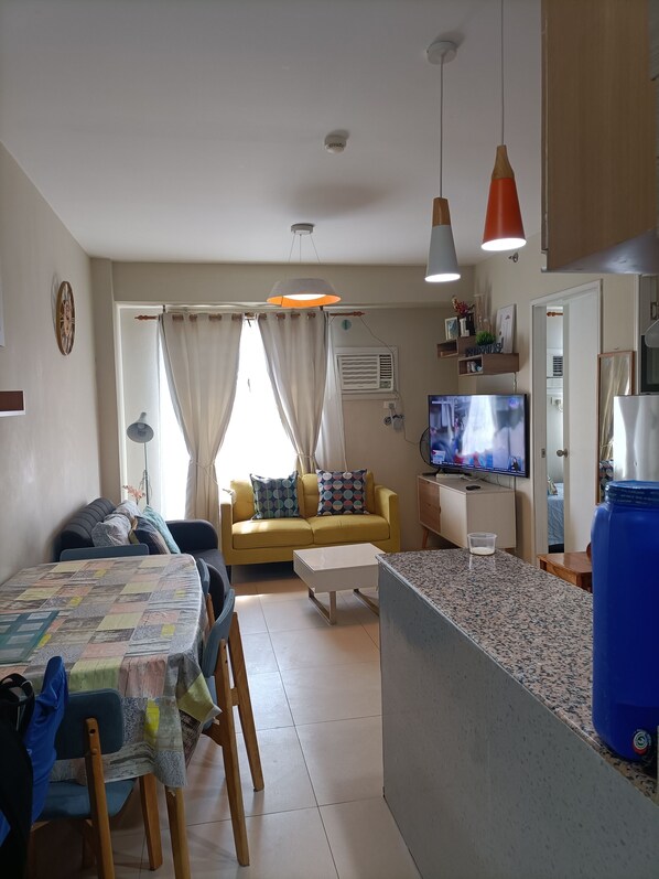 A living room with a yellow sofa, a coffee table, a TV, and a kitchen area with a blue water dispenser. - Avida Tower Sucat (Parañaque)