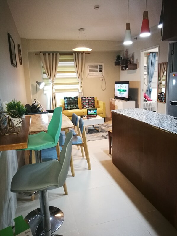 A modern living room with a dining area, a yellow sofa, a TV, and a kitchen area with a refrigerator. - Avida Tower Sucat (Parañaque)