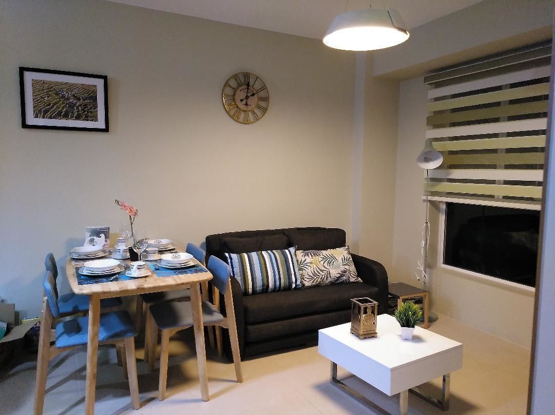 A living room with a black sofa, a wooden dining table set with plates and cups, a white coffee table, and a clock on the wall.
