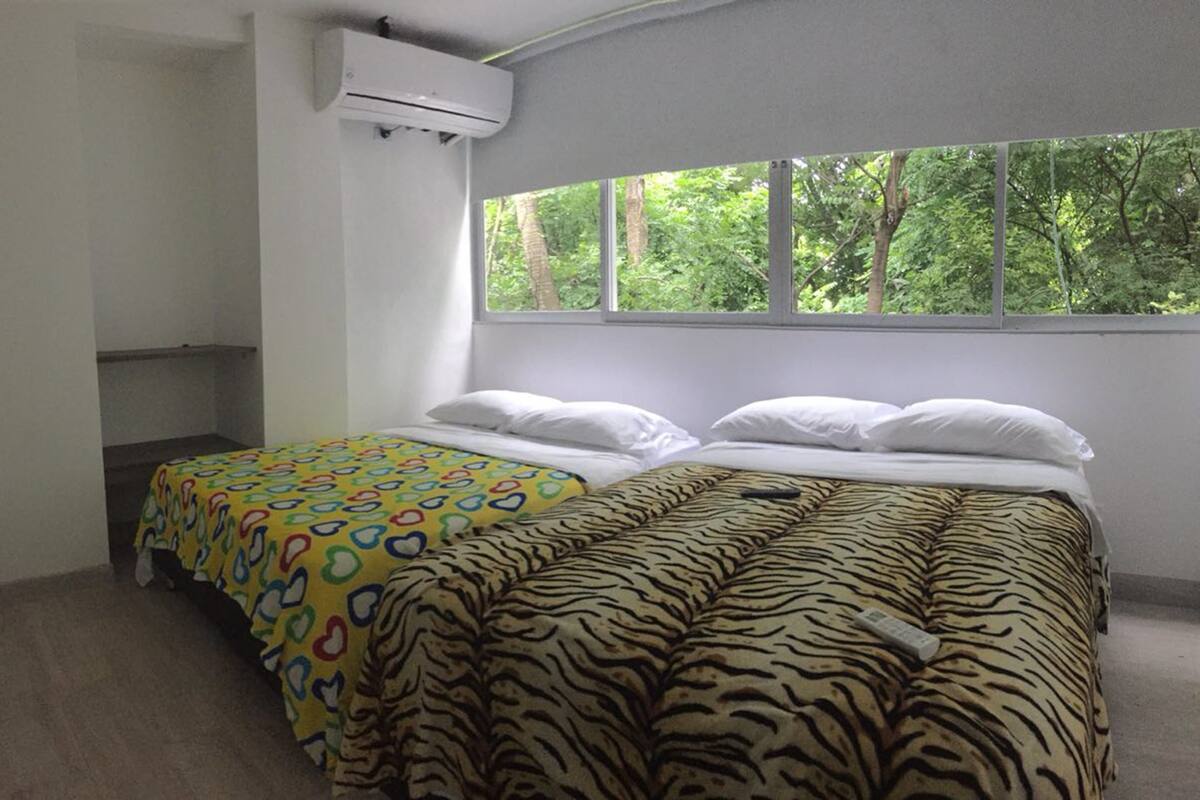 Une chambre avec deux lits, une fenêtre donnant sur les arbres et un appareil de climatisation.