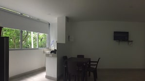 Une pièce moderne et bien éclairée, comprenant un coin repas, une petite cuisine et une grande fen être donnant sur la verdure.