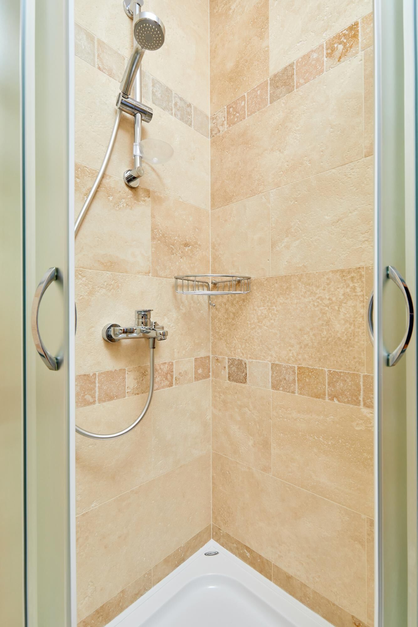 Un espace douche doté d’une porte en verre, d’un pommeau de douche à main, d’un robinet mural et d’une petite étagère.