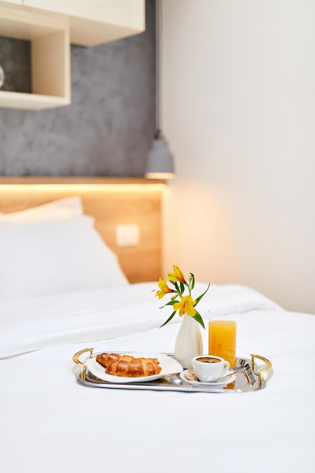 A neatly made breakfast tray with croissants, coffee, orange juice, and a vase of flowers on a hotel bed.