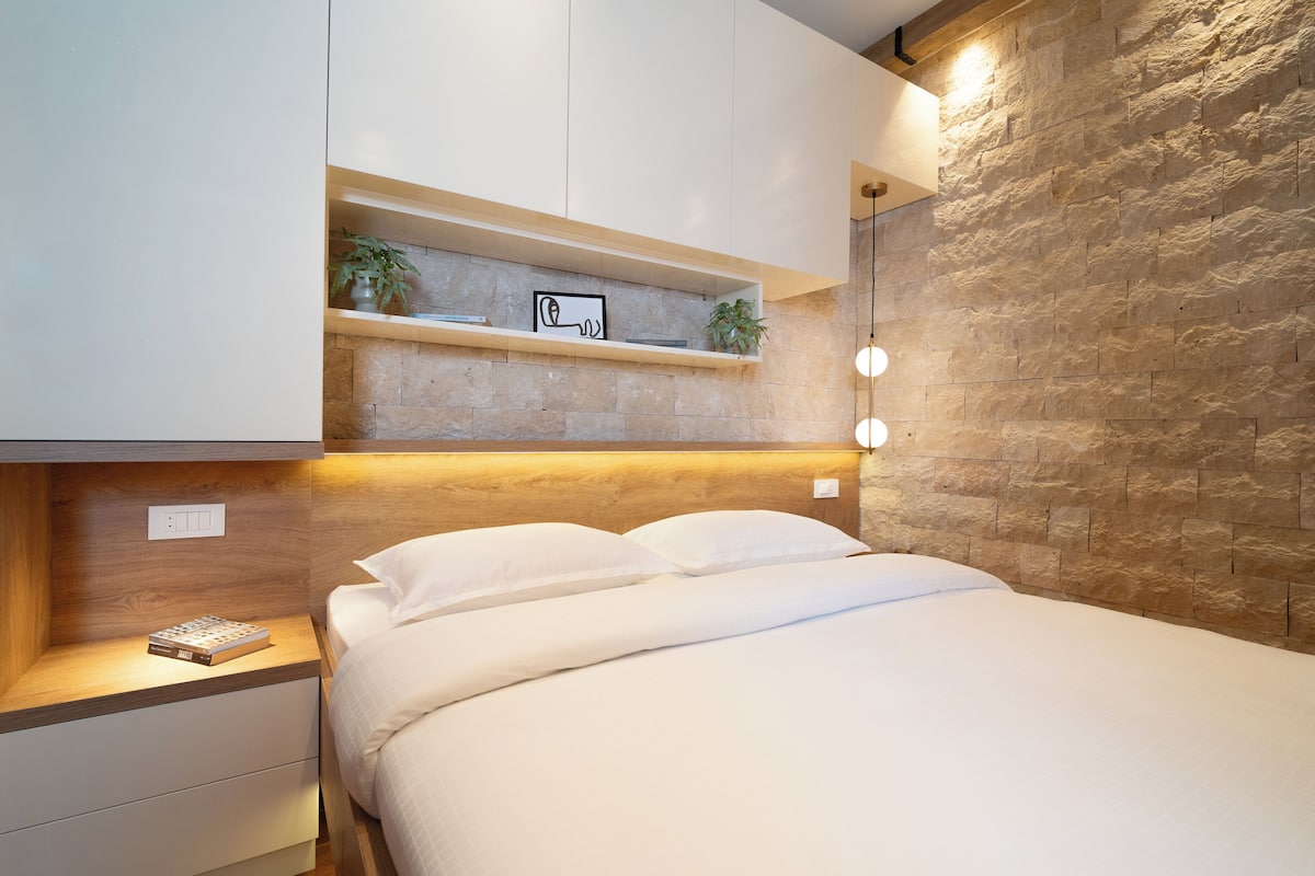 A modern bedroom with a stone accent wall, a bedside table with a book, and a shelf with decorative items.