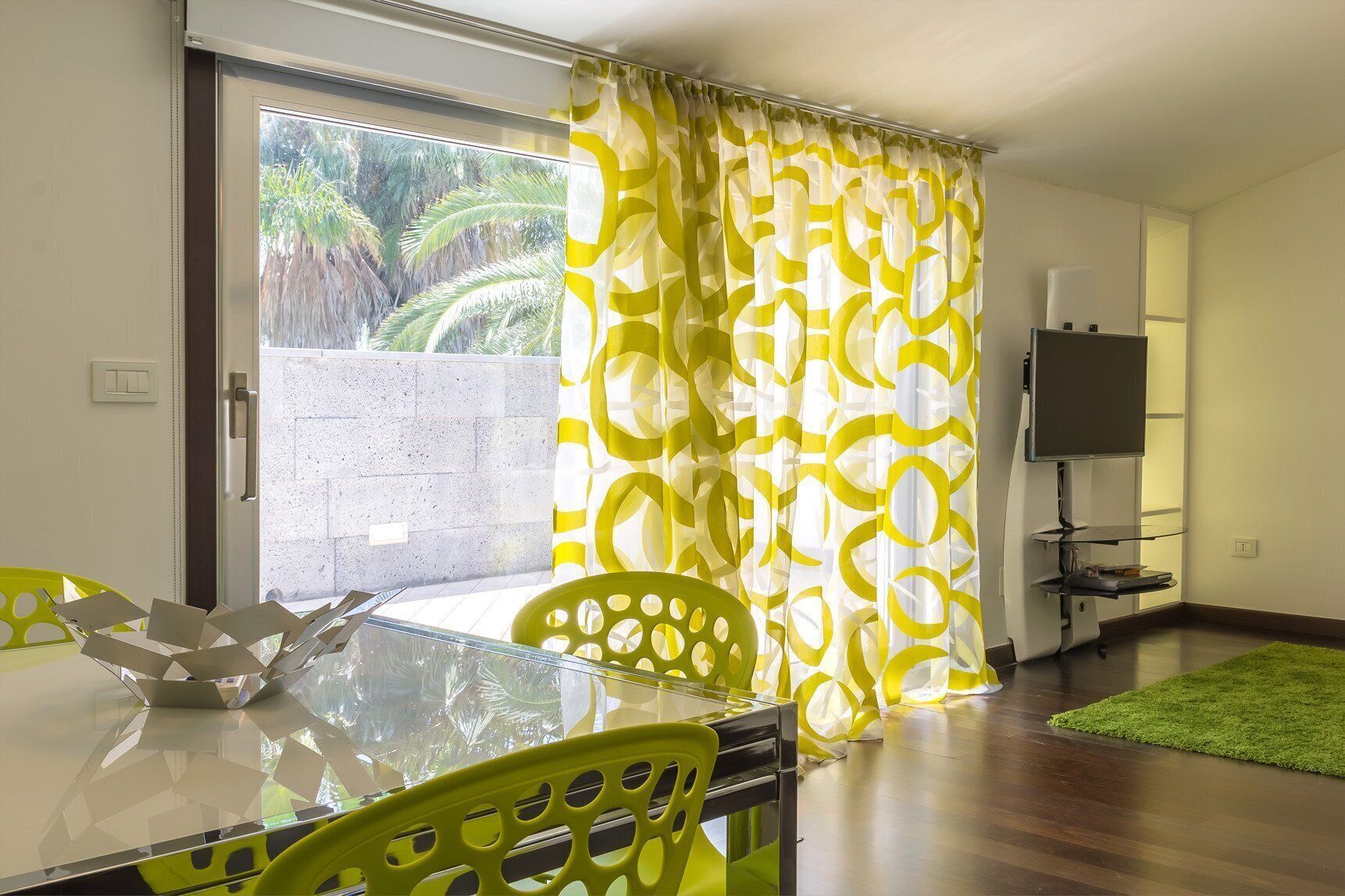 A dining area with a glass table, yellow chairs, a TV on a stand, and a view of palm trees through the window.