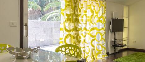A dining area with a glass table, yellow chairs, a TV on a stand, and a view of palm trees through the window.